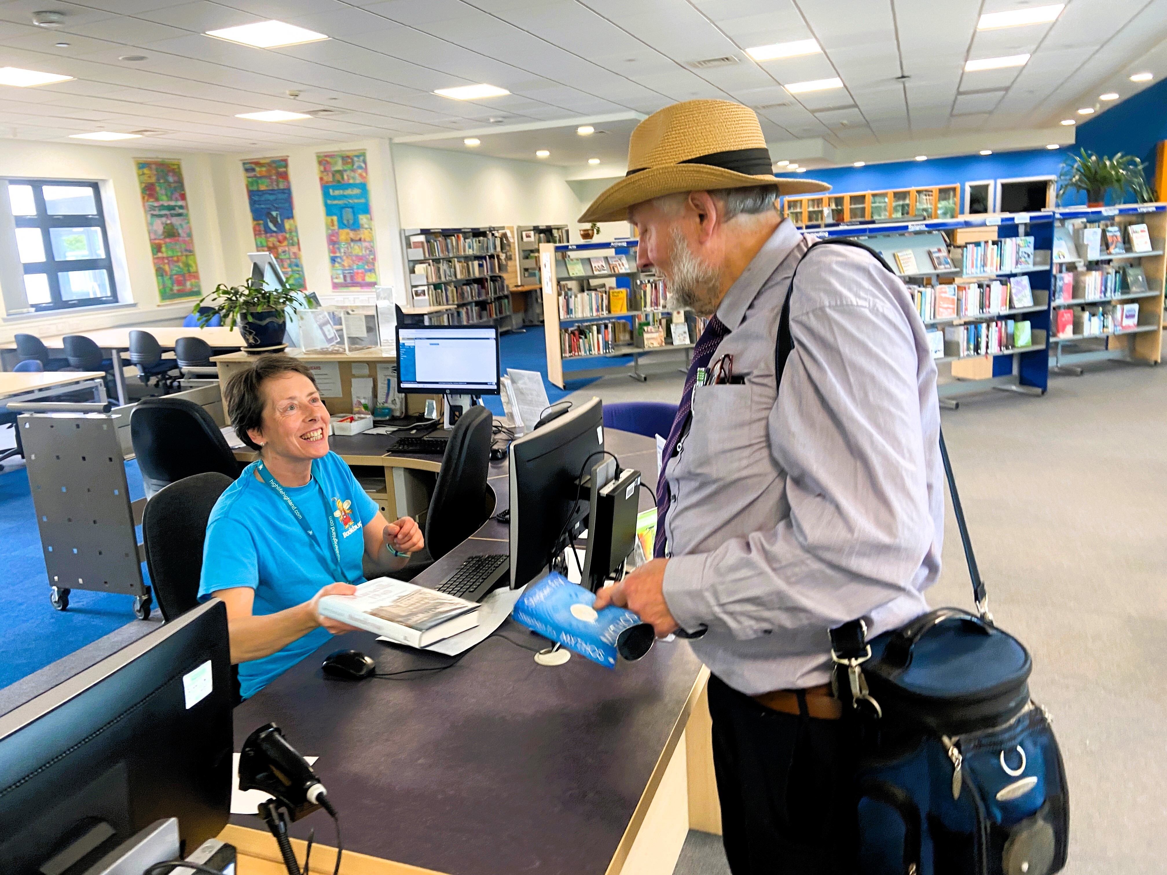 A library staff member helps a visitor at a service desk, with bookshelves and computer areas visible in the bright, open library space.
