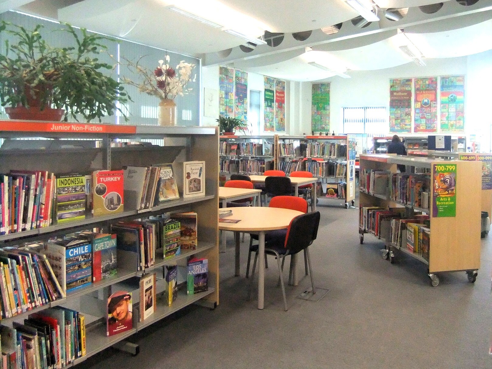 Bright children’s library with shelves of colourful books, round tables with chairs, and posters on the walls in a spacious, well‑lit room.