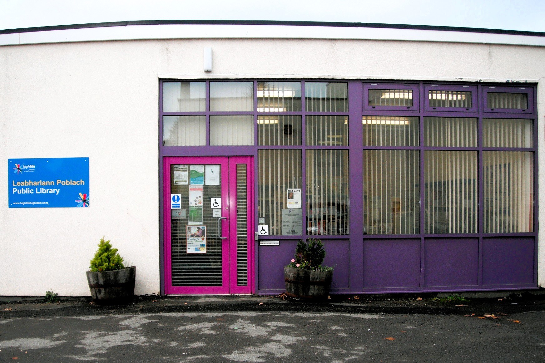 Entrance of Golspie Library, white building with large purple-framed windows and a blue bilingual sign.
