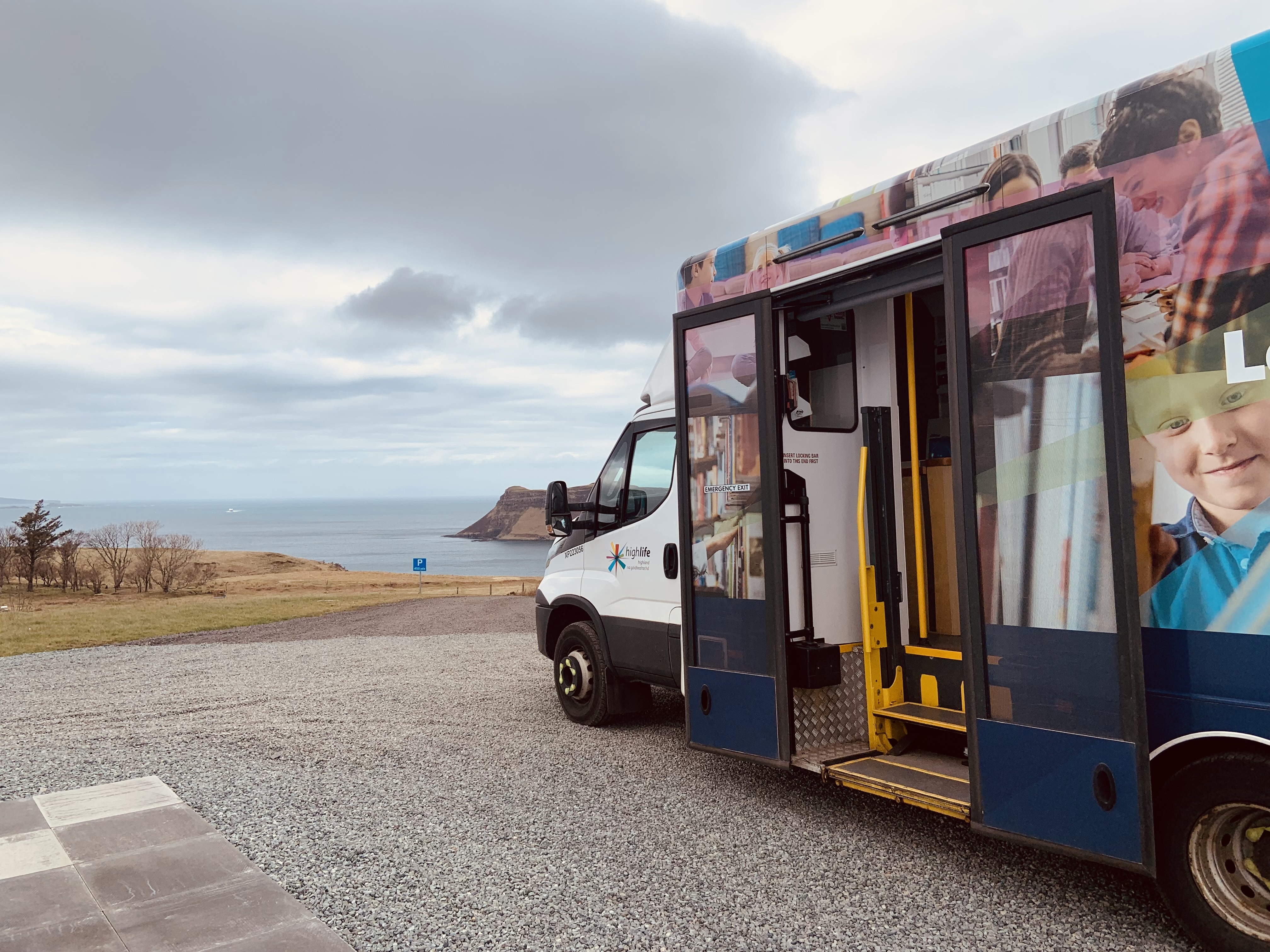 A mobile library van parked on a gravel area overlooking a coastal landscape, with the sea, cliffs, and cloudy sky in the background. The van’s door is open, showing the entrance steps and part of the interior.