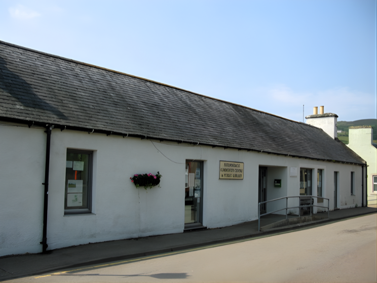 A long white single‑storey library building with a slate roof, central glass door, side ramp entrance and a hanging flower basket.
