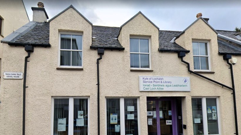 Front of Kyle of Lochalsh Library, cream-colored building with slate roof, purple entrance, and bilingual signage.