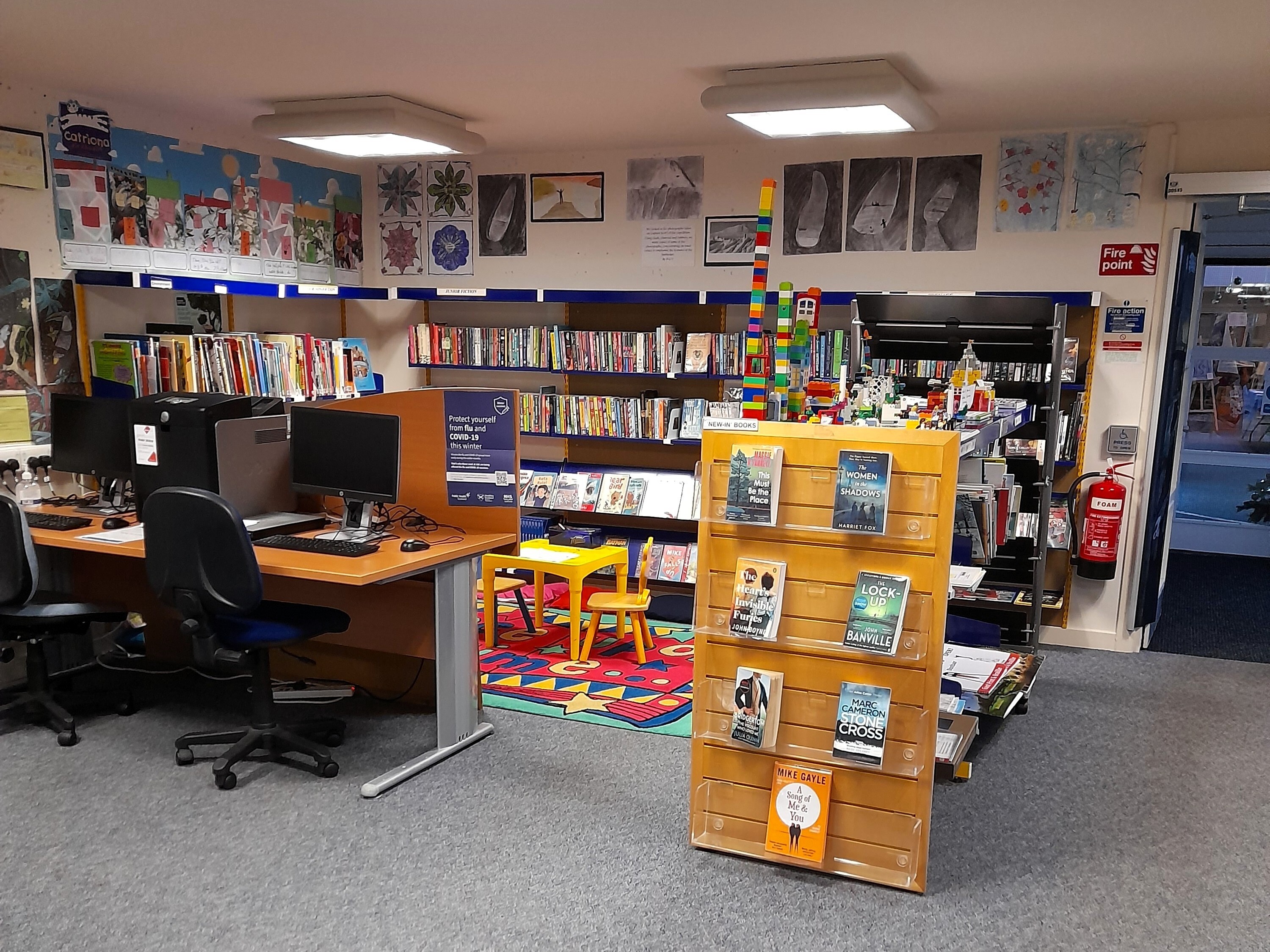 Interior of a small library with public computers on the left, children’s bookshelves and a colourful play mat in the centre, and a display stand of books in the foreground.
