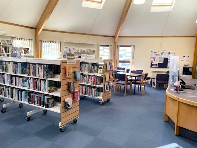 Interior of Mallaig Library with bookshelves, reading table, notice board, and a curved wooden desk under skylights.