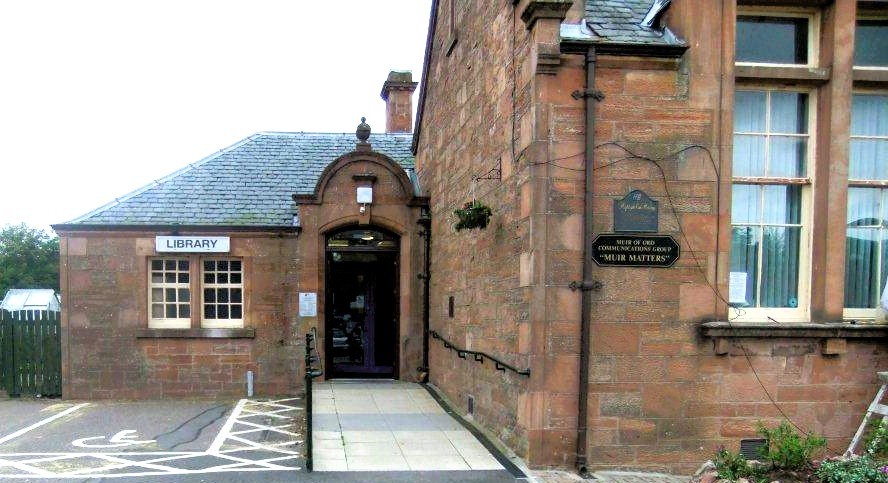 Entrance of Muir of Ord Library, stone building with arched doorway, small windows, and a sign reading ‘Library".