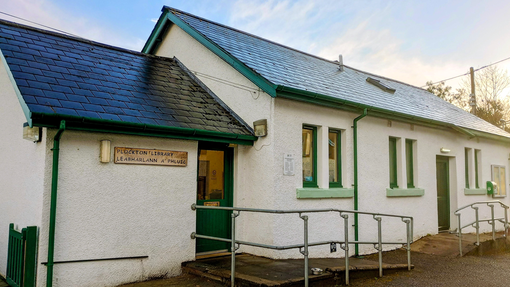 Single‑storey white building with green trim and a sign reading ‘Plockton Library’. A ramp with railings leads to the entrance.