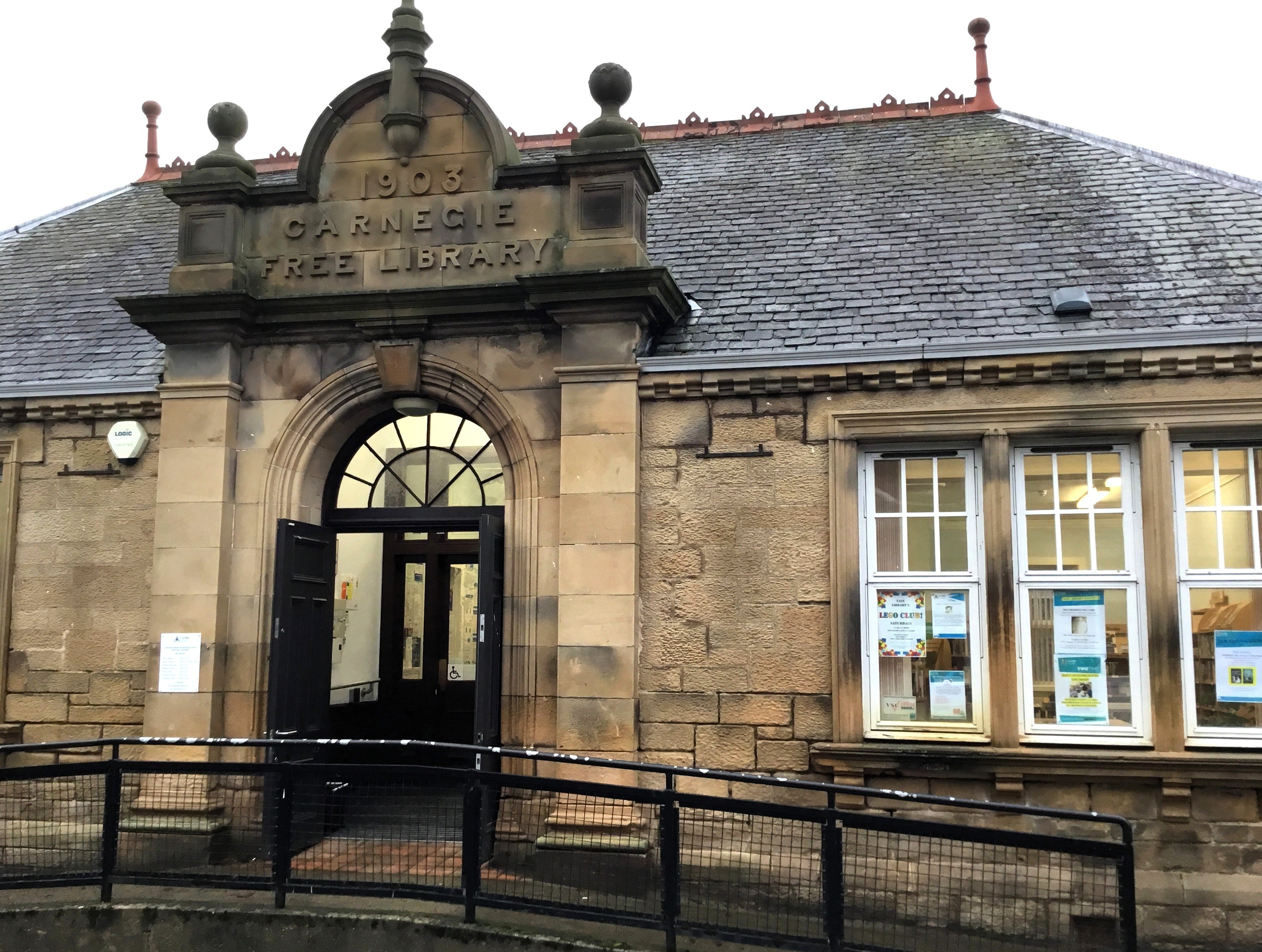 Front of Tain Library, historic stone building with arched doorway, large windows, and ‘Carnegie Free Library’ inscription.