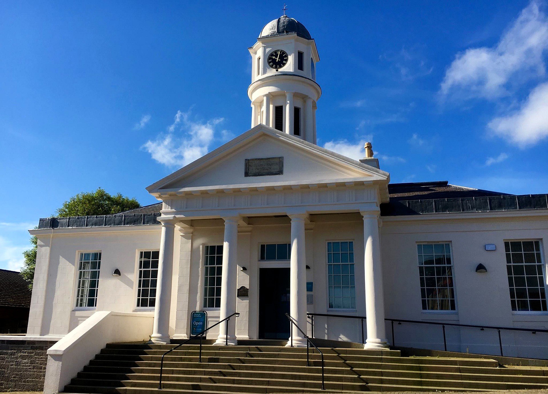 White neoclassical building with four columns at the entrance, wide steps leading up, and a clock tower with a domed roof against a bright blue sky.