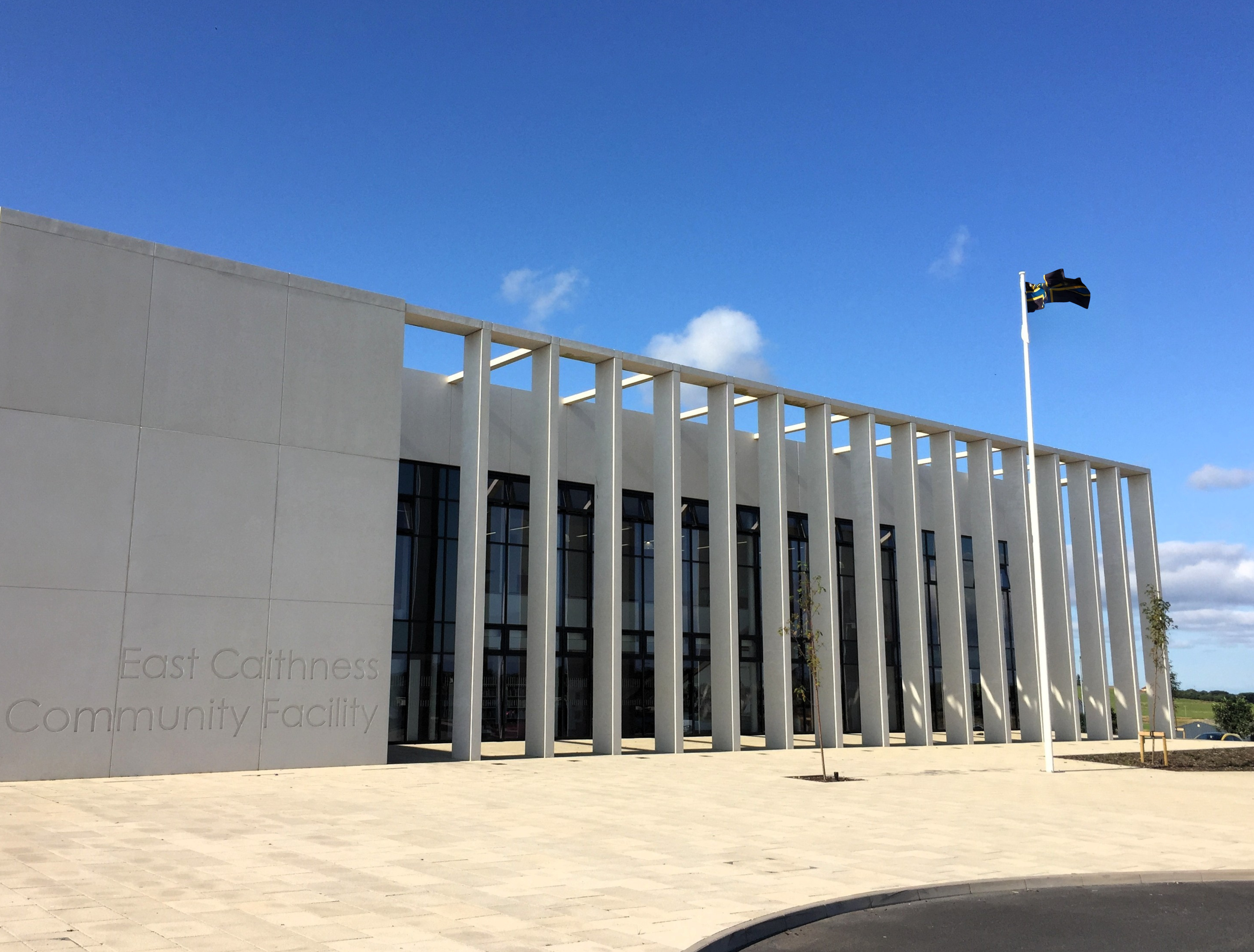 Modern white building with tall vertical columns, large glass windows, and a flagpole in front under a clear blue sky.