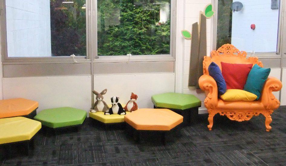 Bright reading corner with colorful hexagonal stools, plush toys, and an ornate orange armchair with multicolored cushions near large windows.