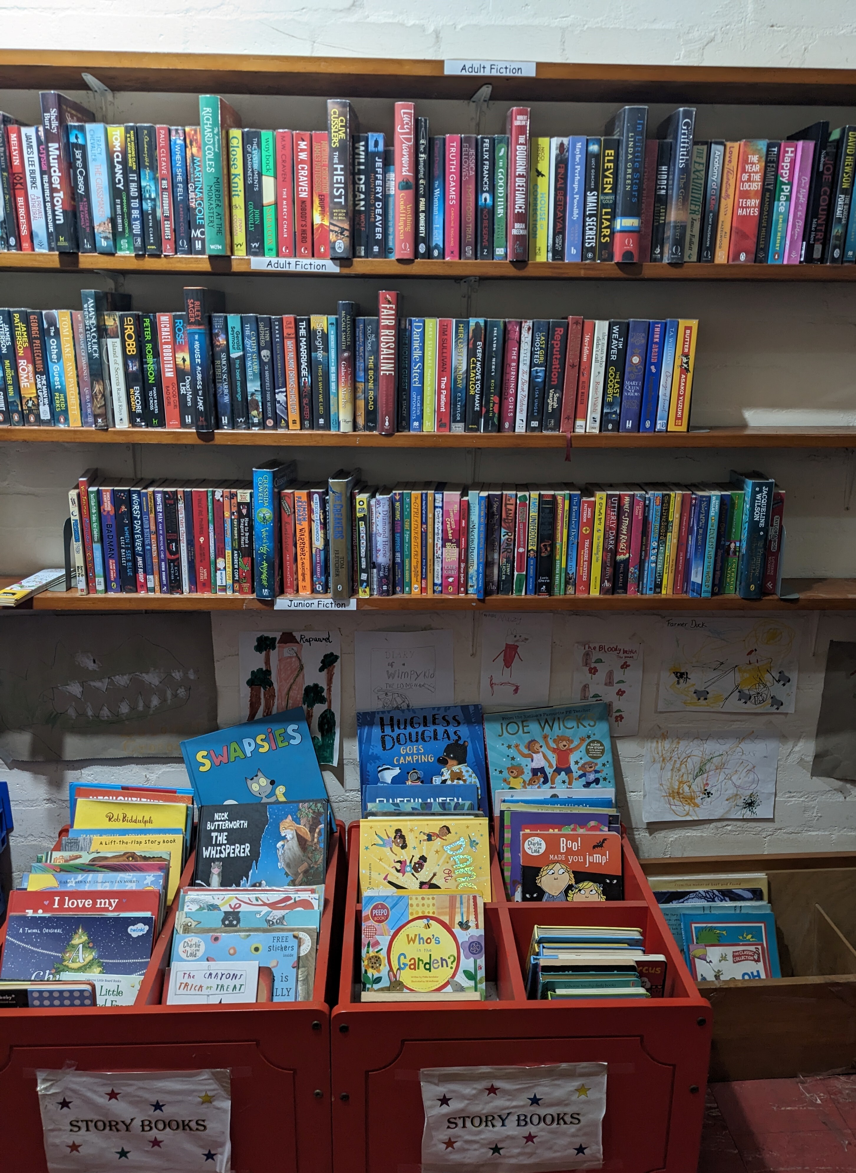 Library shelves with adult fiction above and children’s picture books in red bins below, with kids’ drawings on the wall behind.