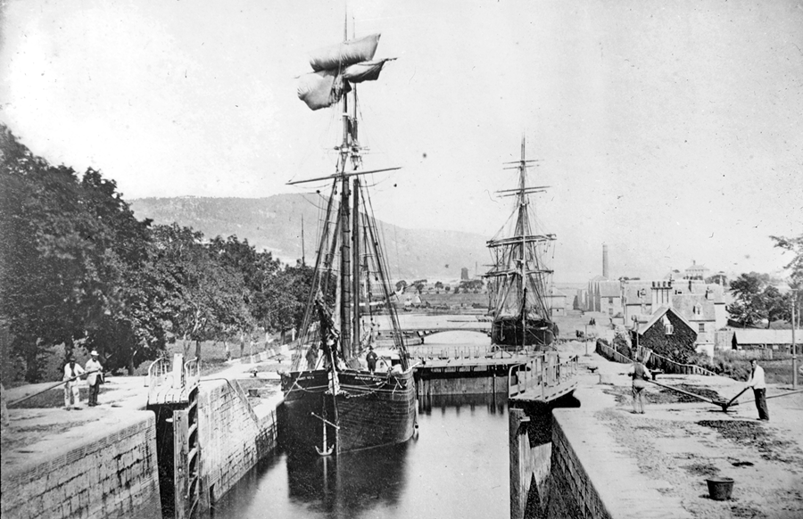 A historic photograph of two sailing ships passing through a canal lock, with people standing along the lock walls and buildings and hills visible in the background.