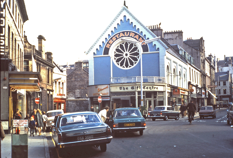 A street scene shows the historic stained glass Rose Window with cars and people in front of a blue building with a large circular window and a sign reading “Restaurant.” Below it is “The Gift Centre.” Surrounding buildings line the street on both sides.