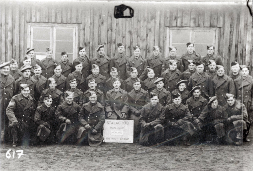 A black‑and‑white group photograph shows rows of uniformed individuals posed outside a wooden building. A sign in the front row reads “STALAG XVII B — Main Camp — Wolk K.C. — District Group.” The number “617” appears in the lower left corner. The image shows signs of age and wear.