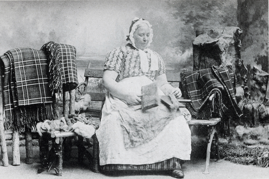 A vintage studio photograph shows a person seated on a bench carding wool, with woven textiles draped over the bench and wool bundles on the ground. The image is labeled “An Cardadh.”
