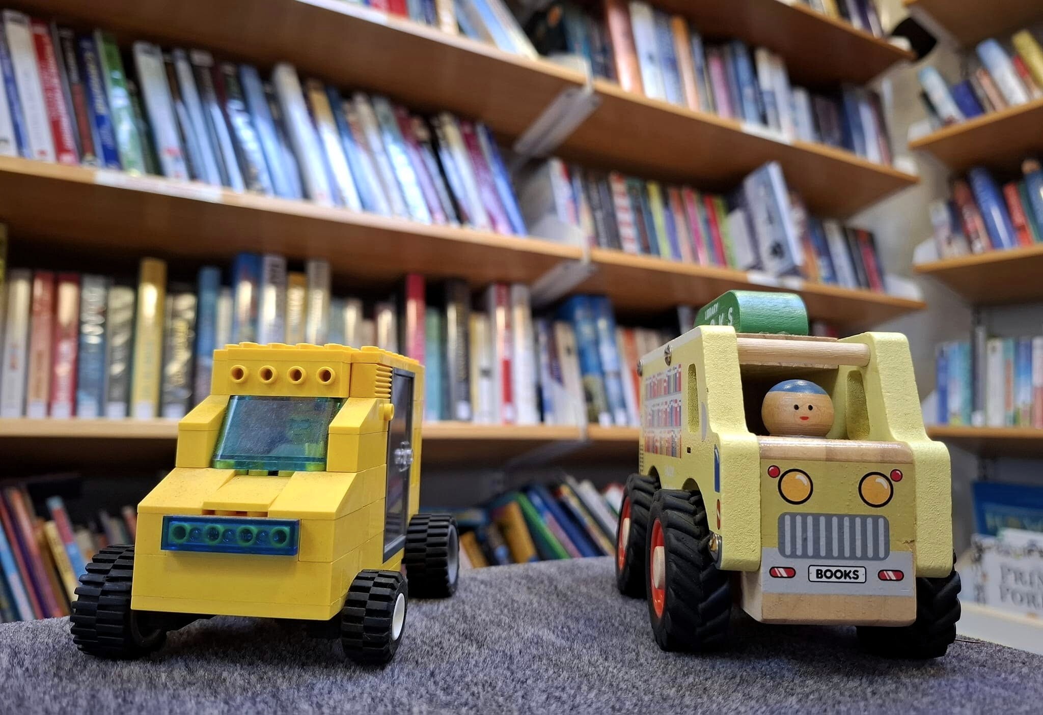 Two small toy vehicles displayed on a surface inside a library. One toy is a yellow block‑style vehicle with chunky wheels. The other is a wooden toy van with illustrated details and a small round‑headed figure sitting inside. Behind them are several shelves filled with books.