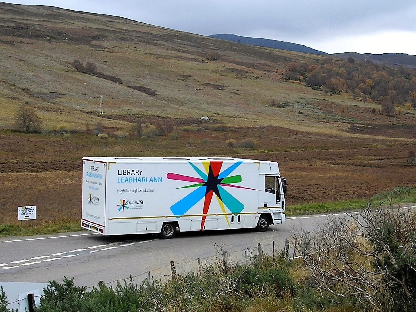 A white mobile library van with a large multicoloured starburst design on its side travels along a rural road. The surrounding landscape consists of rolling brown and green hills with patches of autumn foliage under an overcast sky.
