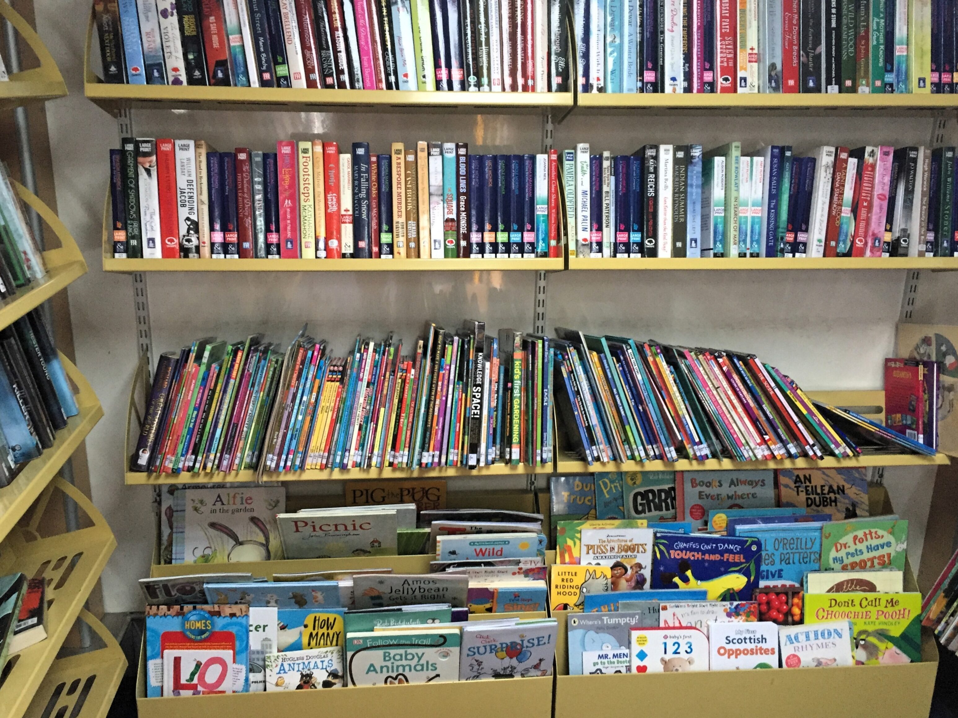 A set of library bookshelves filled with books. The top two shelves hold a variety of adult fiction and non‑fiction books arranged upright. The middle shelf displays children’s picture books and thin illustrated books stacked tightly side‑by‑side. The bottom section contains front‑facing children's books with colourful covers arranged in boxes.