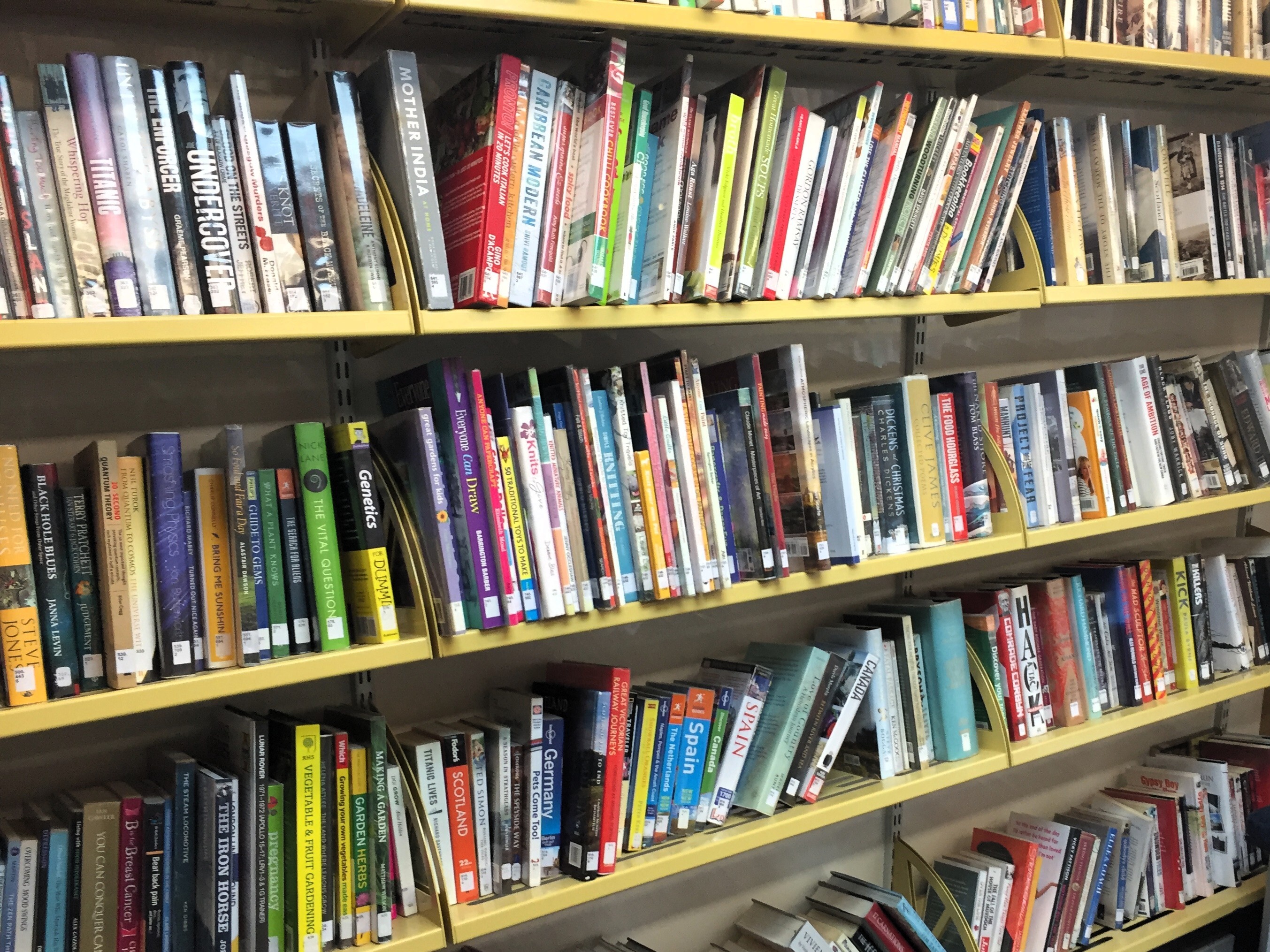 Three long shelves filled with adult non‑fiction and travel books on yellow metal shelving. The books are arranged upright with a mix of colours, sizes, and spine designs. Some titles are grouped by topic, and the shelves are tightly packed across the length of the photo.
