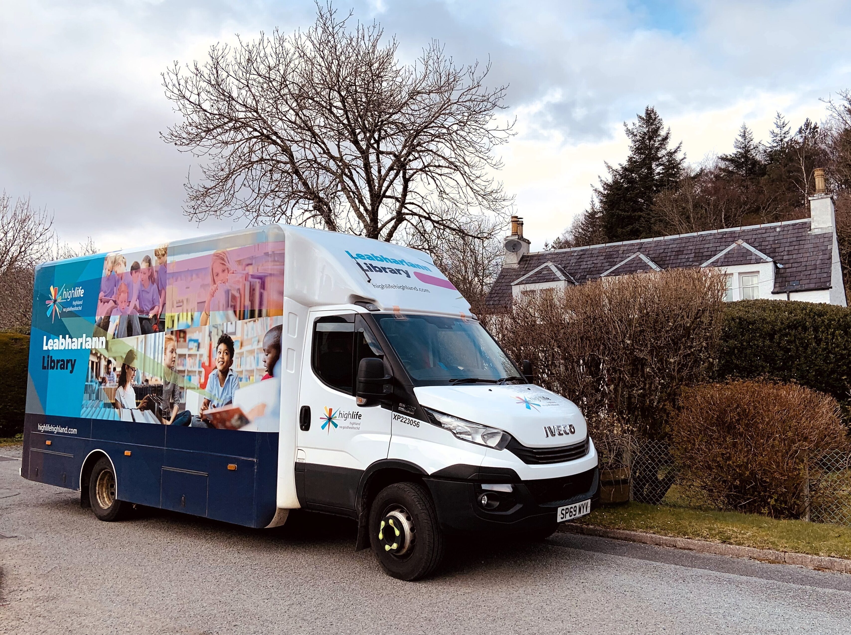 A mobile library van parked on a quiet residential street, with a cottage, trimmed hedges, and leafless trees in the background under a partly cloudy sky.