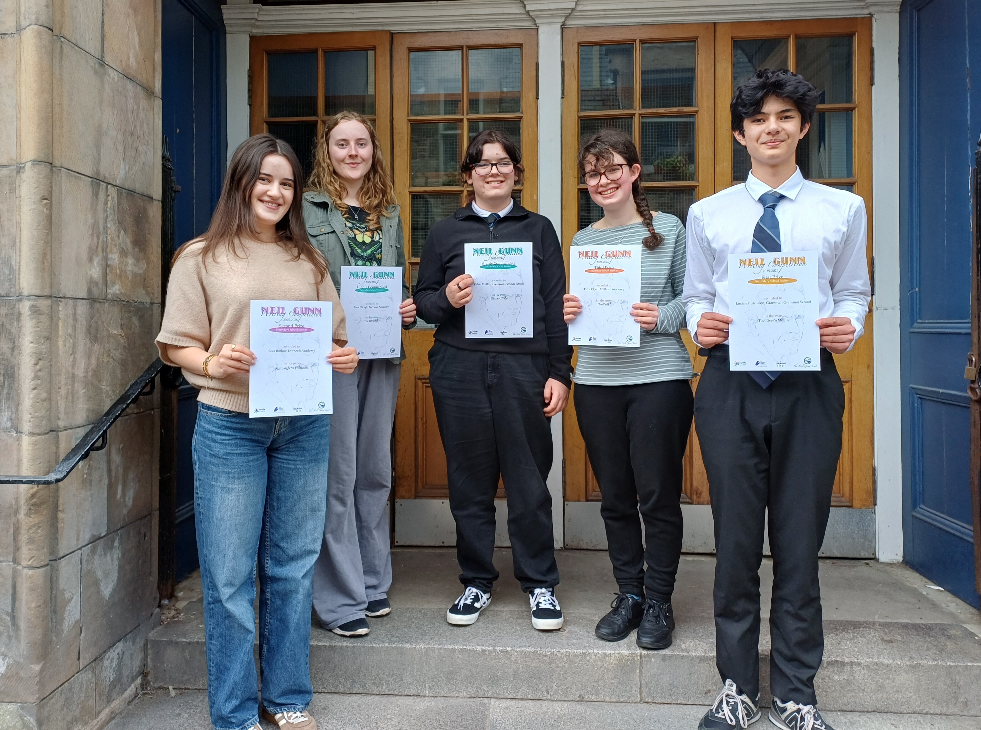 Five students stand on the steps outside a building entrance with wooden double doors, each holding a certificate. The group poses in a row on the stone steps, with the certificates clearly visible, suggesting an awards or achievement presentation. The building has stone walls and blue-painted door frames surrounding the entrance.