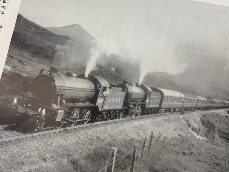 A steam train with two locomotives pulls passenger carriages through hilly countryside, with white steam rising as it travels along a curved track.
