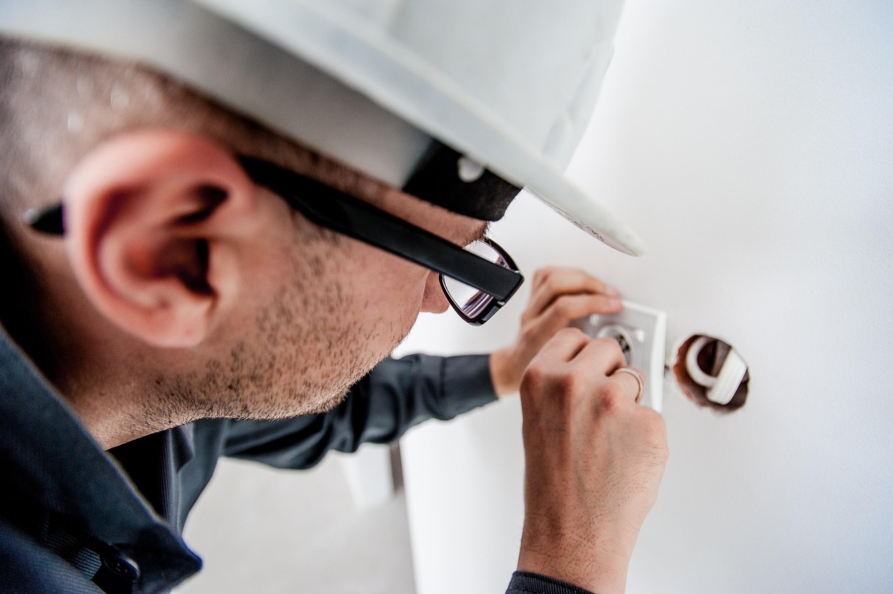 A person wearing a hard hat and glasses is working on an electrical socket in a wall, using a tool to make adjustments. Part of the wiring is visible beside the socket.