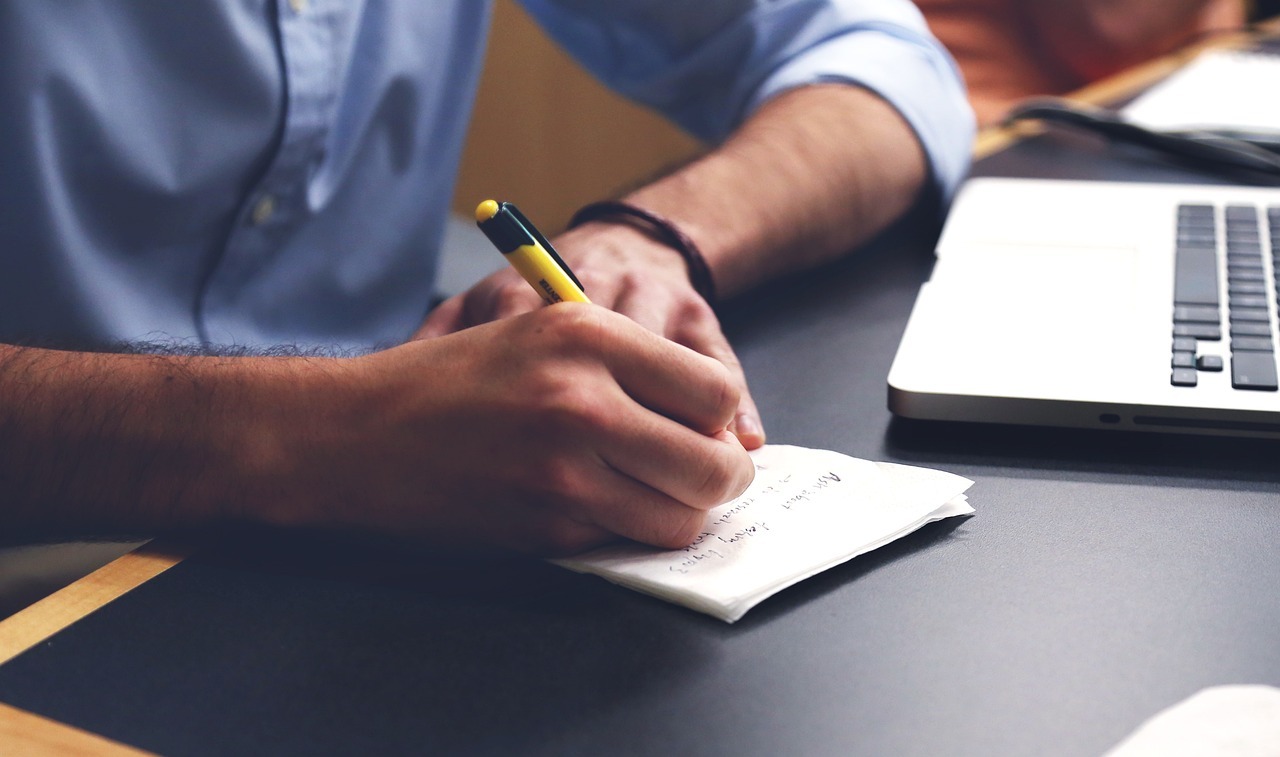 A person seated at a desk writes notes on a small notepad with a pen, beside an open laptop.