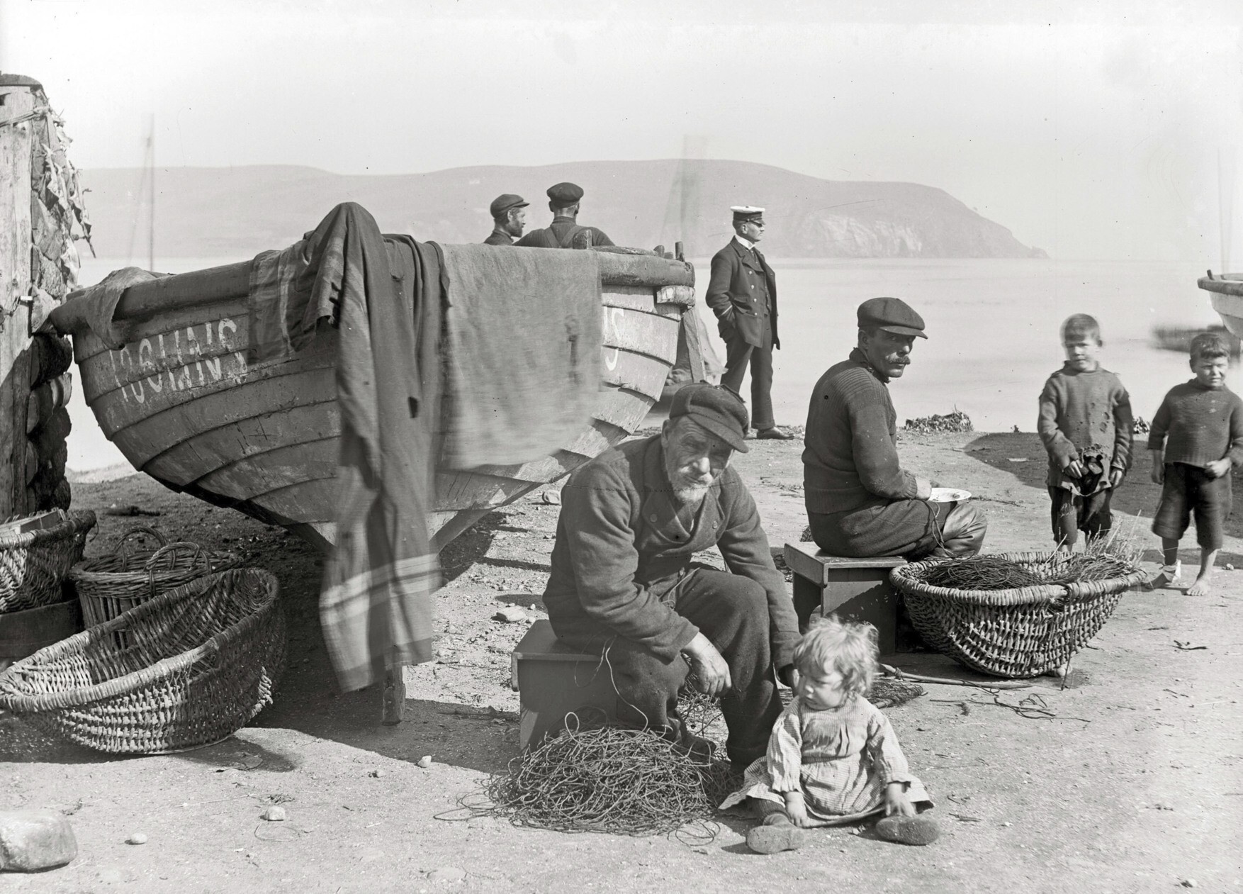 People by a beached boat with baskets and nets on a shoreline, with hills and water in the background and a child sitting on the ground.