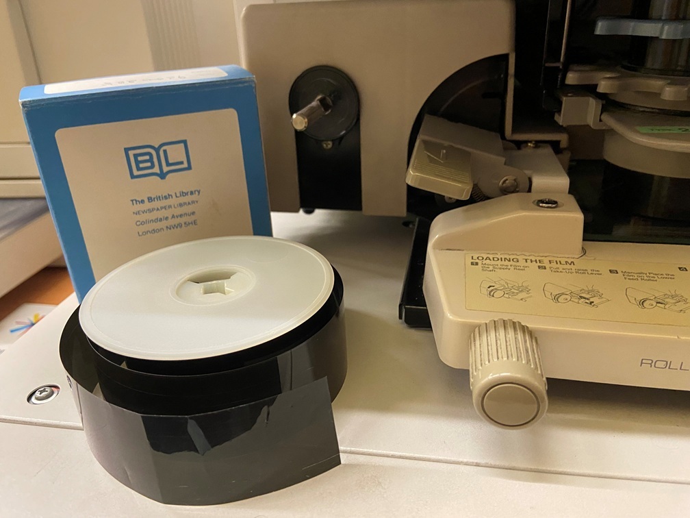 A close-up of a microfilm reel and its storage box beside the loading mechanism of a microfilm reader.