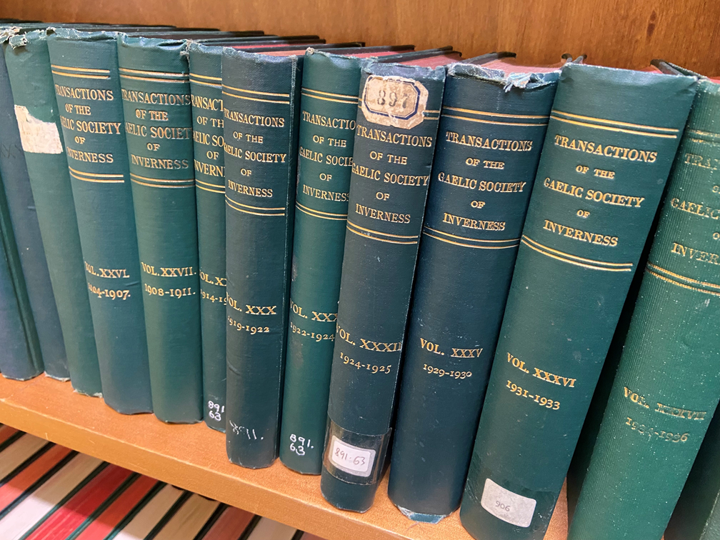 A row of dark green hardback volumes titled ‘Transactions of the Gaelic Society of Inverness’ arranged on a wooden shelf, showing various volumes from the early 1900s.