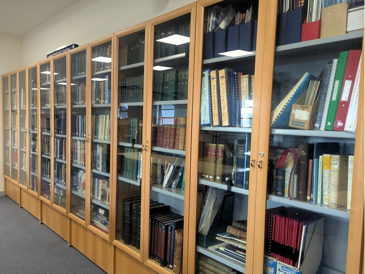 A row of tall wooden library cabinets with glass doors displaying books, folders, and archival materials along a quiet library corridor.