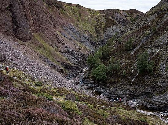 Steep-sided gorge with rocky slopes and patches of heather, leading to a narrow stream at the bottom. A small group of walkers is visible near the water and another person stands on the slope to the left.