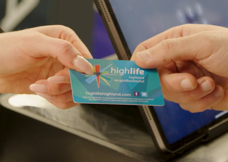 A close-up of two hands exchanging a High Life membership card in front of a touchscreen terminal and card reader at a reception or checkout desk.