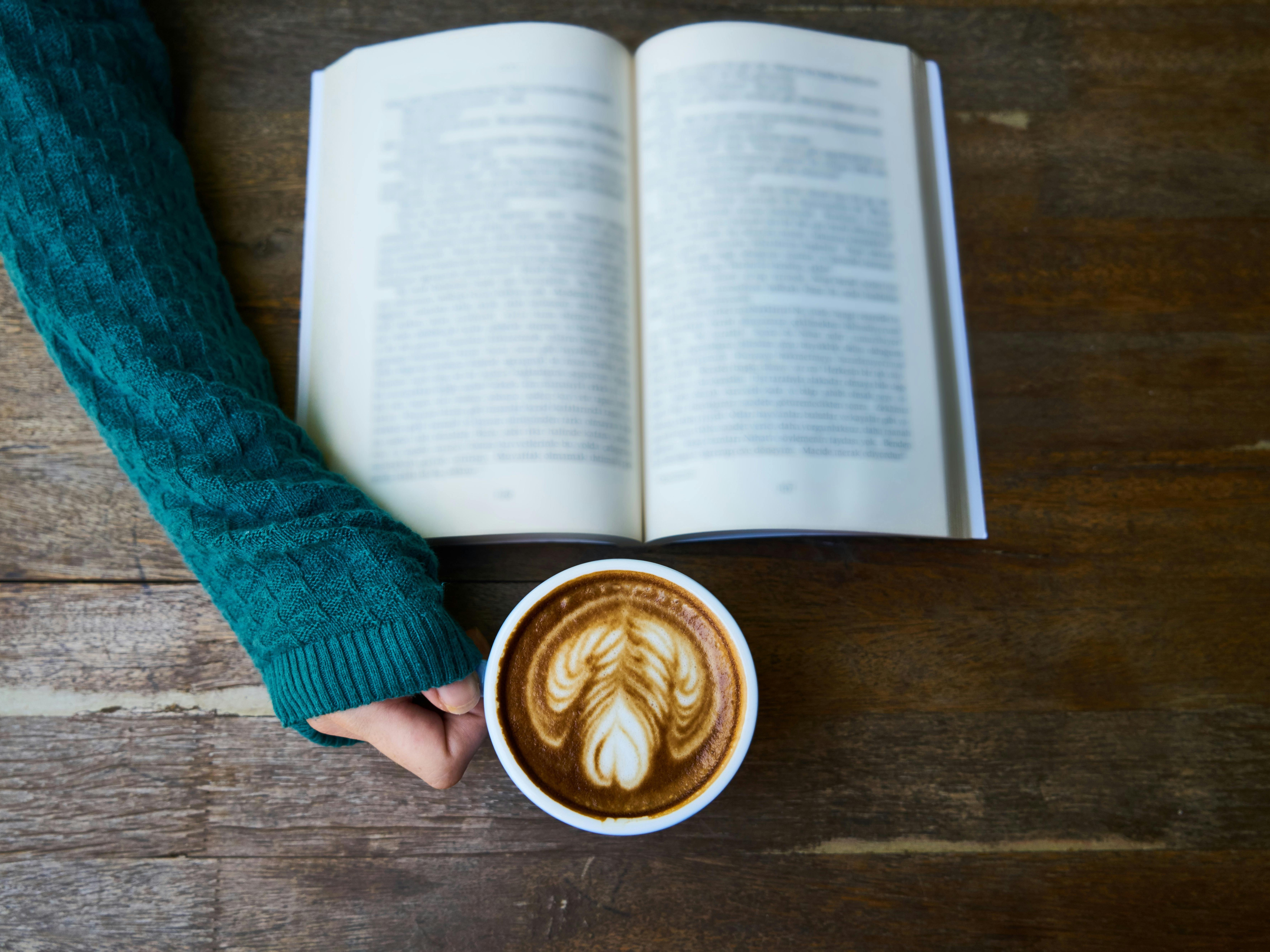 Open book on a wooden table with a person holding a cup of coffee.