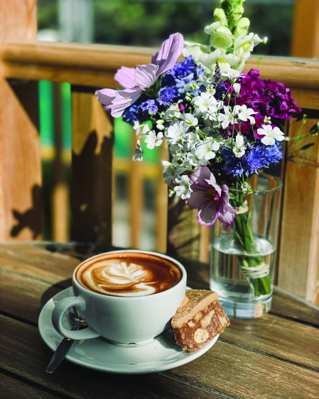 A wooden table at Inverness Botanic Gardens & Café with a white cup of latte featuring leaf-shaped latte art, placed on a saucer with a silver spoon and a piece of nutty biscuit. Beside the cup is a clear glass vase holding a colorful bouquet of fresh flowers, including purple, white, and pink blooms. Sunlight casts soft shadows across the rustic wooden surface, creating a warm and inviting atmosphere.