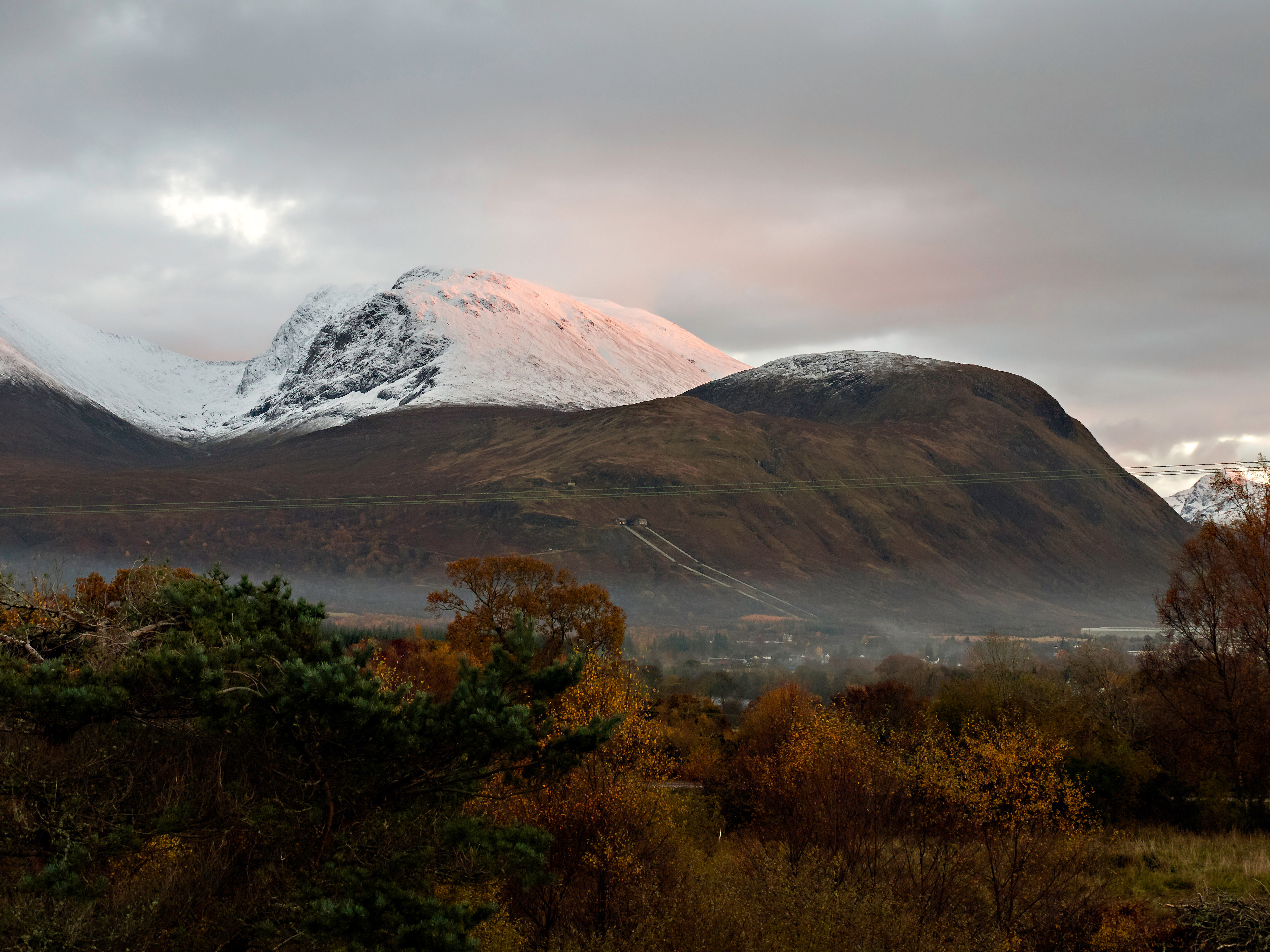 Snow-capped Ben Nevis under a dramatic cloudy sky, with soft pink light from the setting or rising sun illuminating the peak. The foreground features autumnal trees in shades of orange and green, and a misty layer near the base of the mountain adds depth to the scene. The rugged slopes of the mountain contrast with the vibrant valley below.