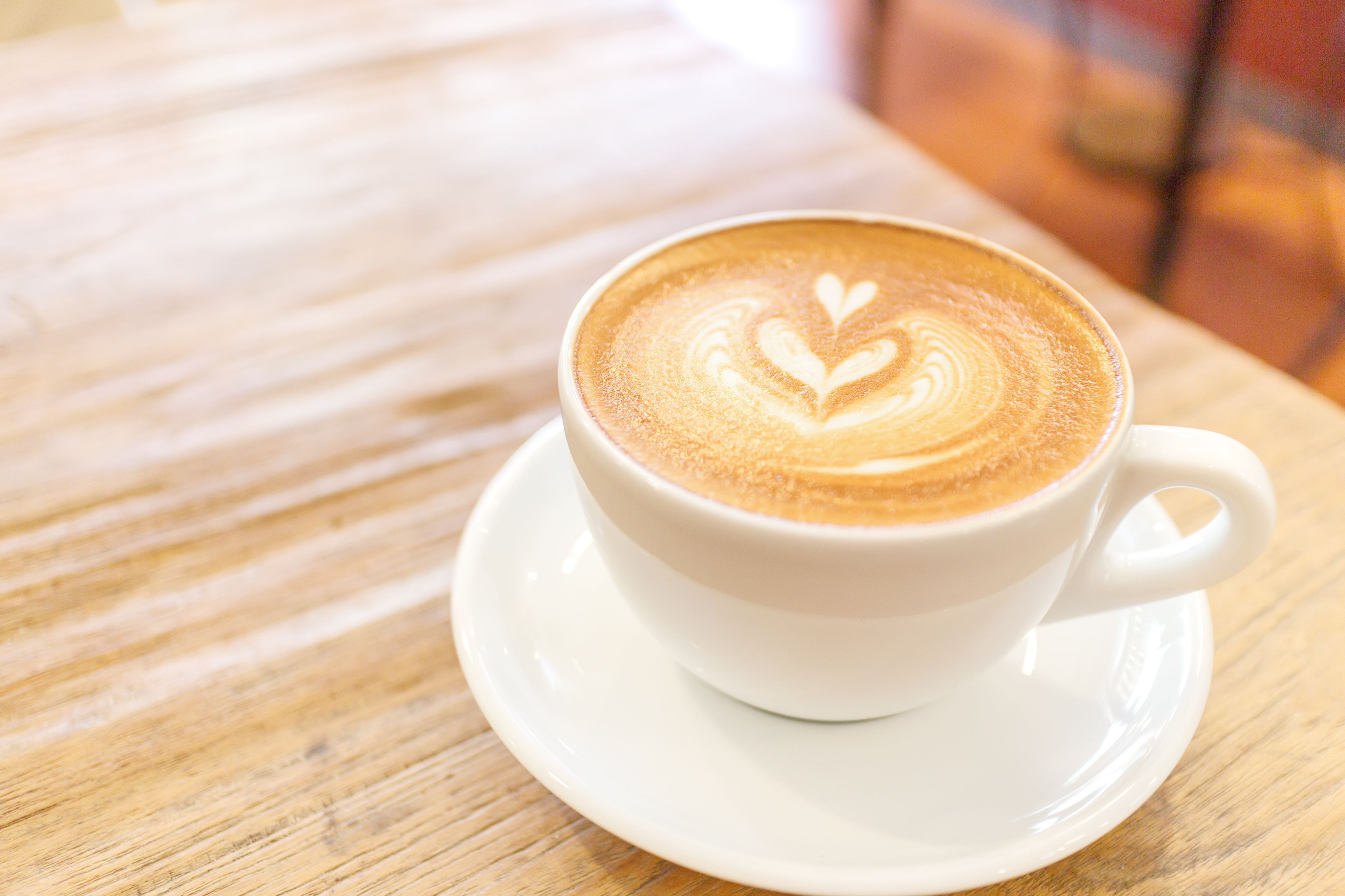 A white ceramic cup filled with a latte featuring heart-shaped latte art on top, placed on a matching saucer on a light wooden table. The background is softly blurred, showing warm tones that suggest an indoor café setting.