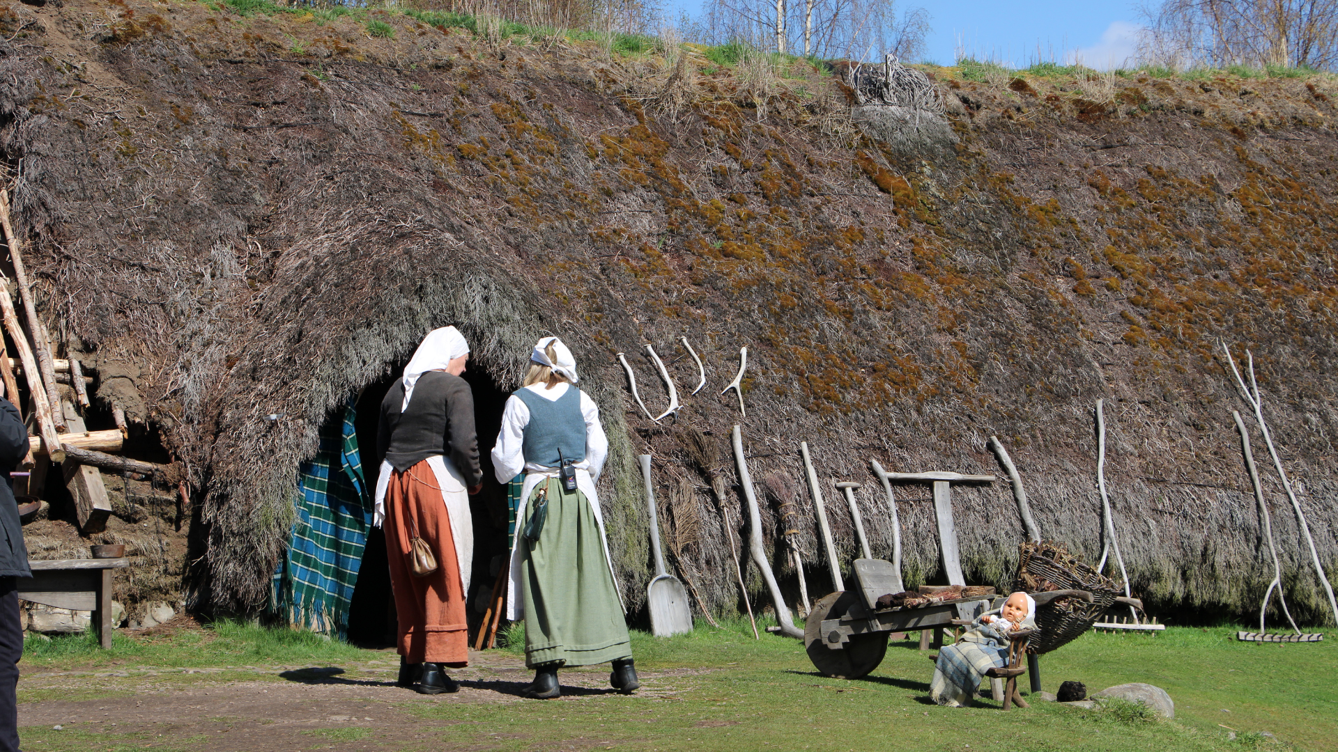 Two people dressed in traditional historical clothing stand outside a thatched-roof building at the Highland Folk Museum. The structure is covered in moss and grass, with wooden farming tools such as rakes and shovels leaning against the wall. A small wooden cart and two dolls in period attire are placed on the ground nearby, with logs stacked to the left and trees visible in the background under a clear blue sky.