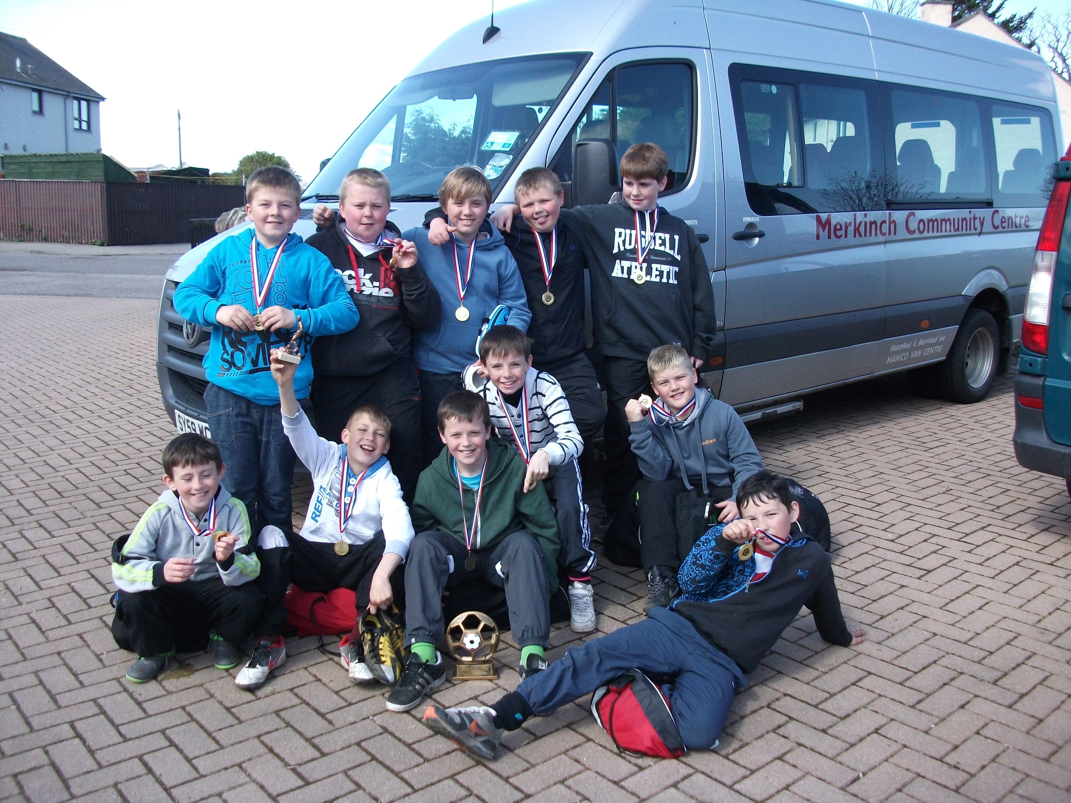 A group of eleven young boys are standing and sitting in front of a silver mini bus. They are all holding up medals and smiling and there is a trophy on the ground.