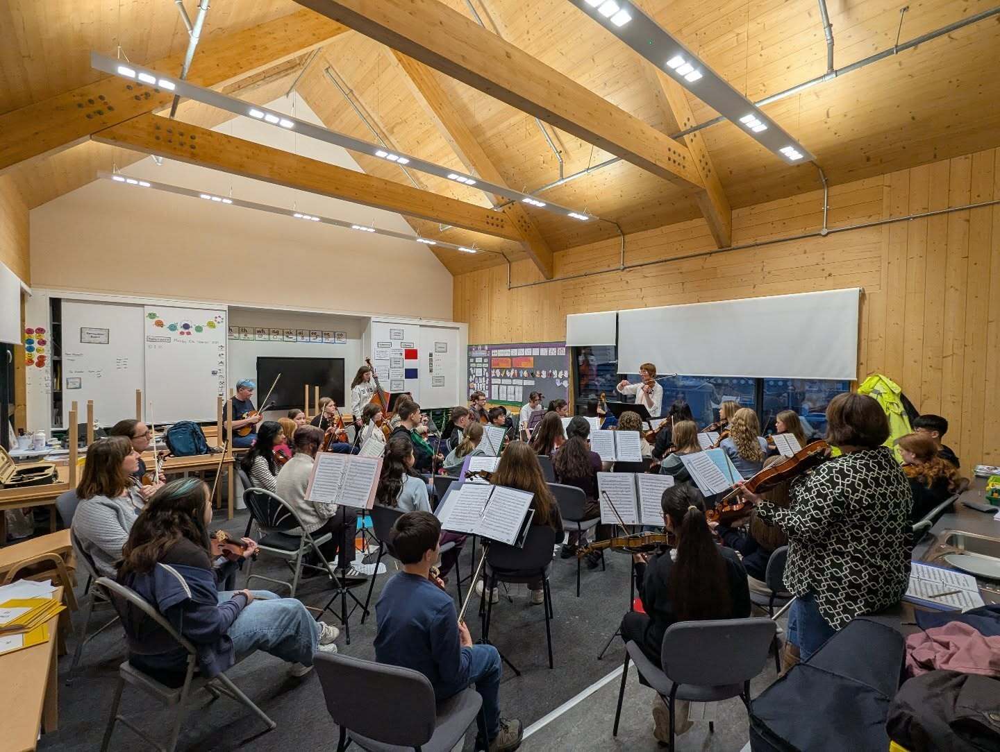 Large group of pupils rehearsing with string instruments in a school music room.