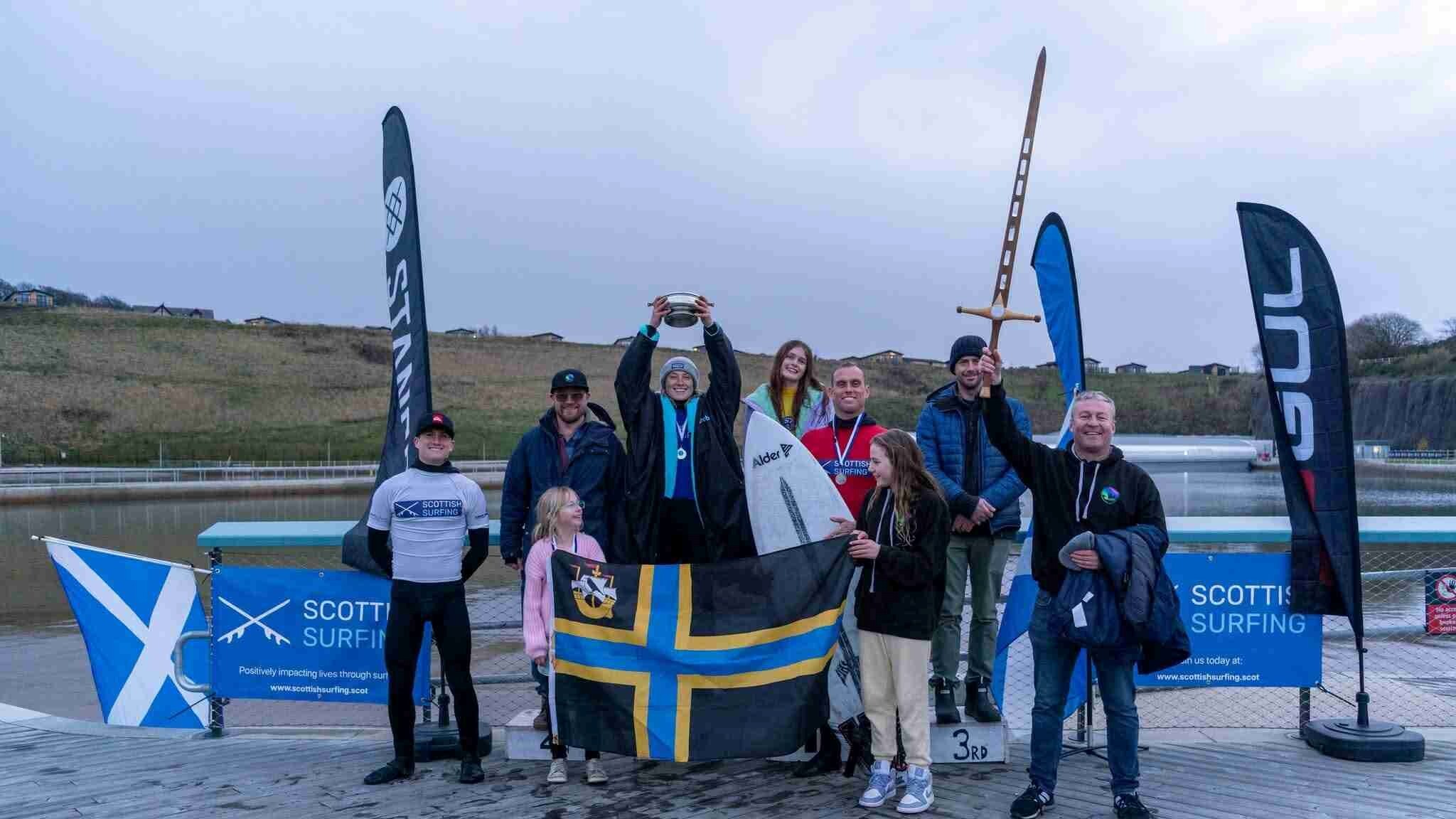 A group of nine surfers are standing on a pier with the sea and hills behind them. There are various flags and banners and one person is holding a sword and another is holding a trophy in the air. There is also another person holding a white surfboard.
