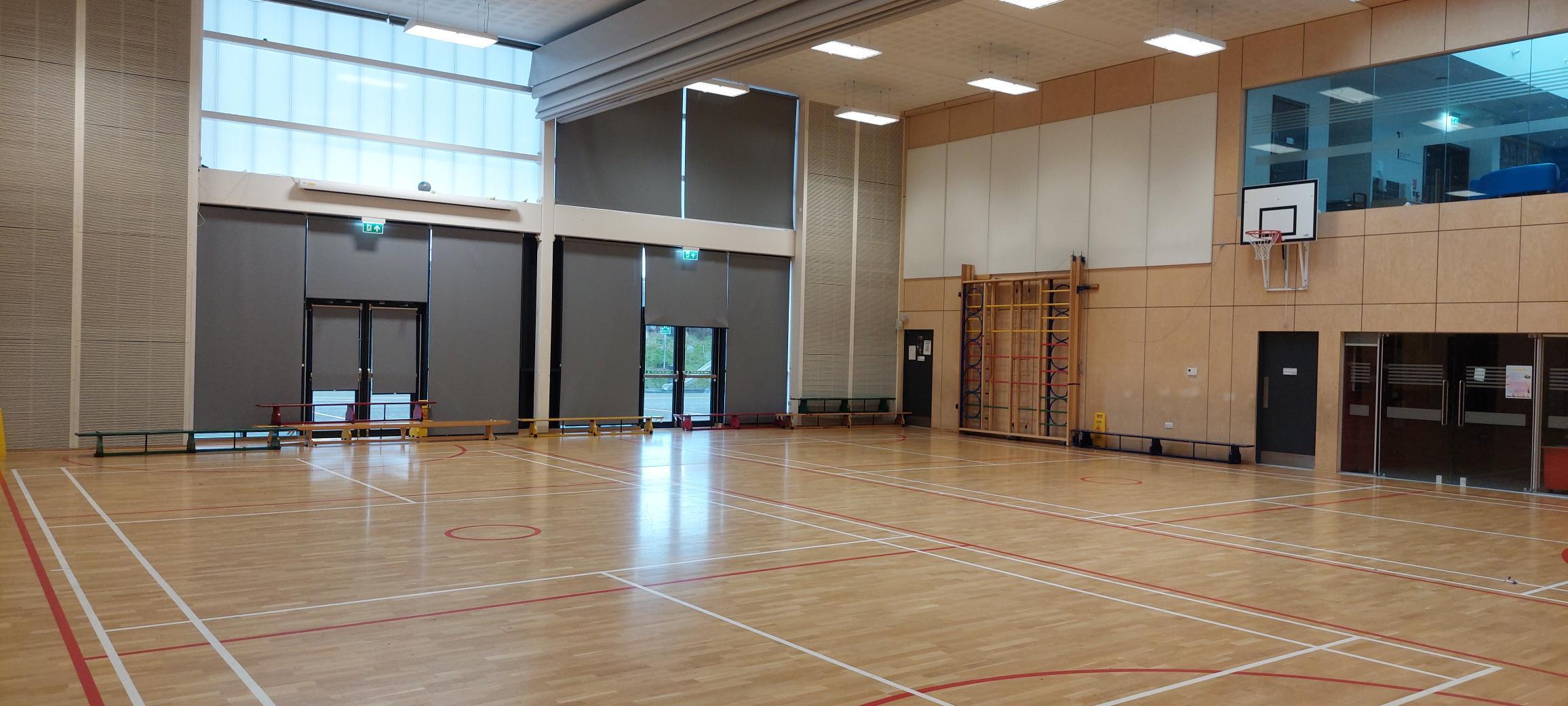 An inside view of a gymnasium hall with court lines for badminton and netball plus basketball hoops mounted to the wall.