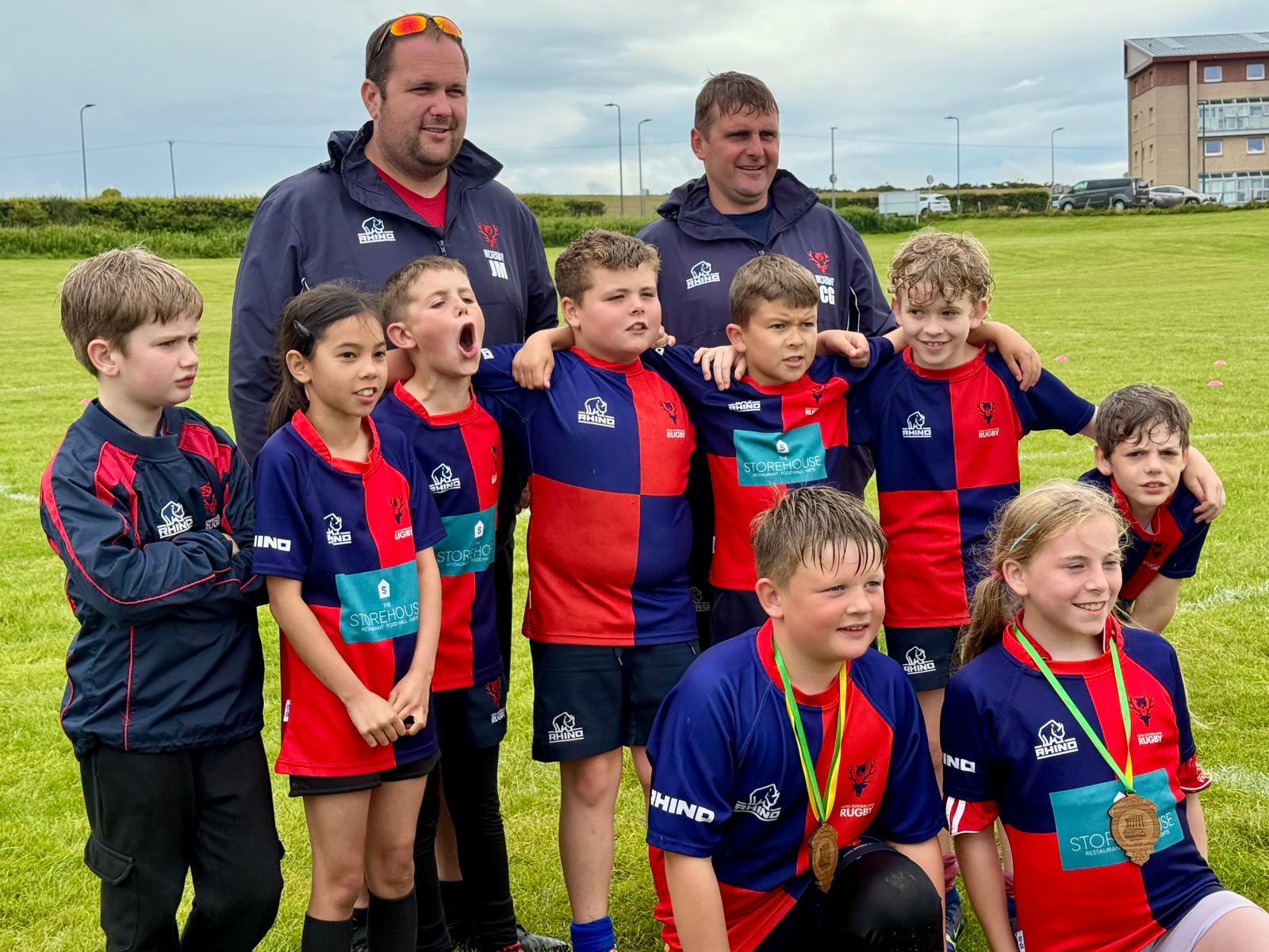 Nine young rugby players are standing in front of two adult male coaches on a sports pitch. The children are wearing blue and red rugby tops and the coaches are wearing blue jumpers. Two of the children at the front are wearing medals.