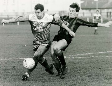 A black‑and‑white photograph of two football players competing for the ball during a match. One player in a light‑coloured kit is kicking the ball while another player in a dark striped kit challenges closely from the side. Both are mid‑stride on a grass pitch with buildings visible in the background.