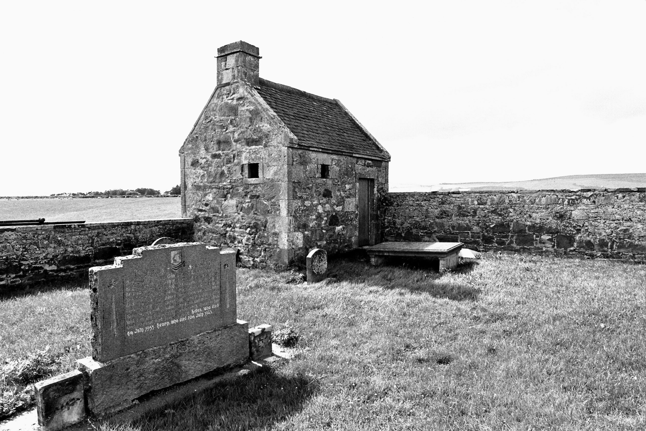 Black-and-white photograph of a small stone watch house within a walled enclosure, surrounded by grass. In the foreground, there is a large upright gravestone with inscriptions and a flat stone tomb nearby. The background shows open countryside beyond the stone wall.