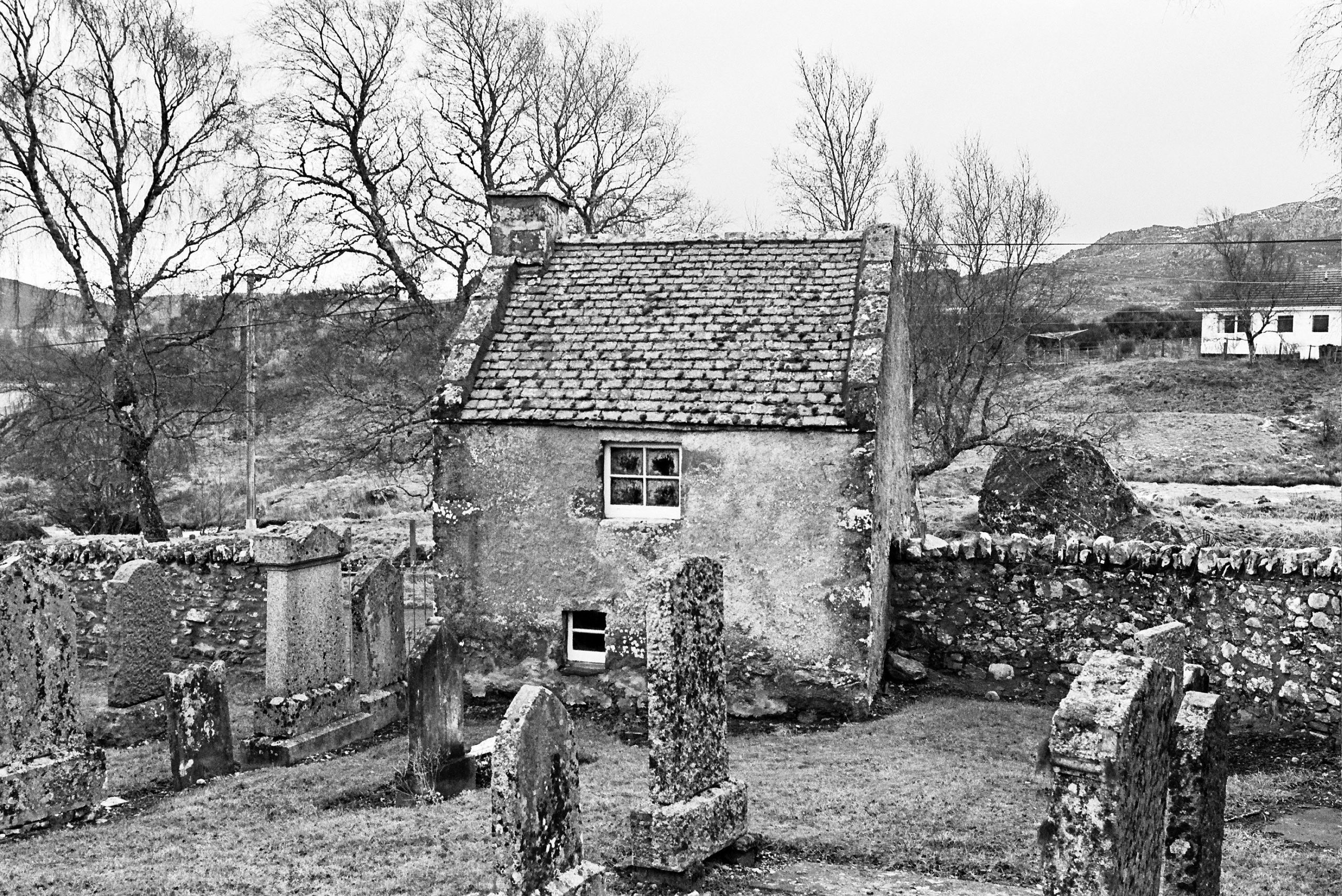 Black-and-white photograph of a small stone watch house with a tiled roof and two windows, set within a walled graveyard. Several weathered gravestones stand in the foreground, and bare trees and hills are visible in the background, along with a distant white building.