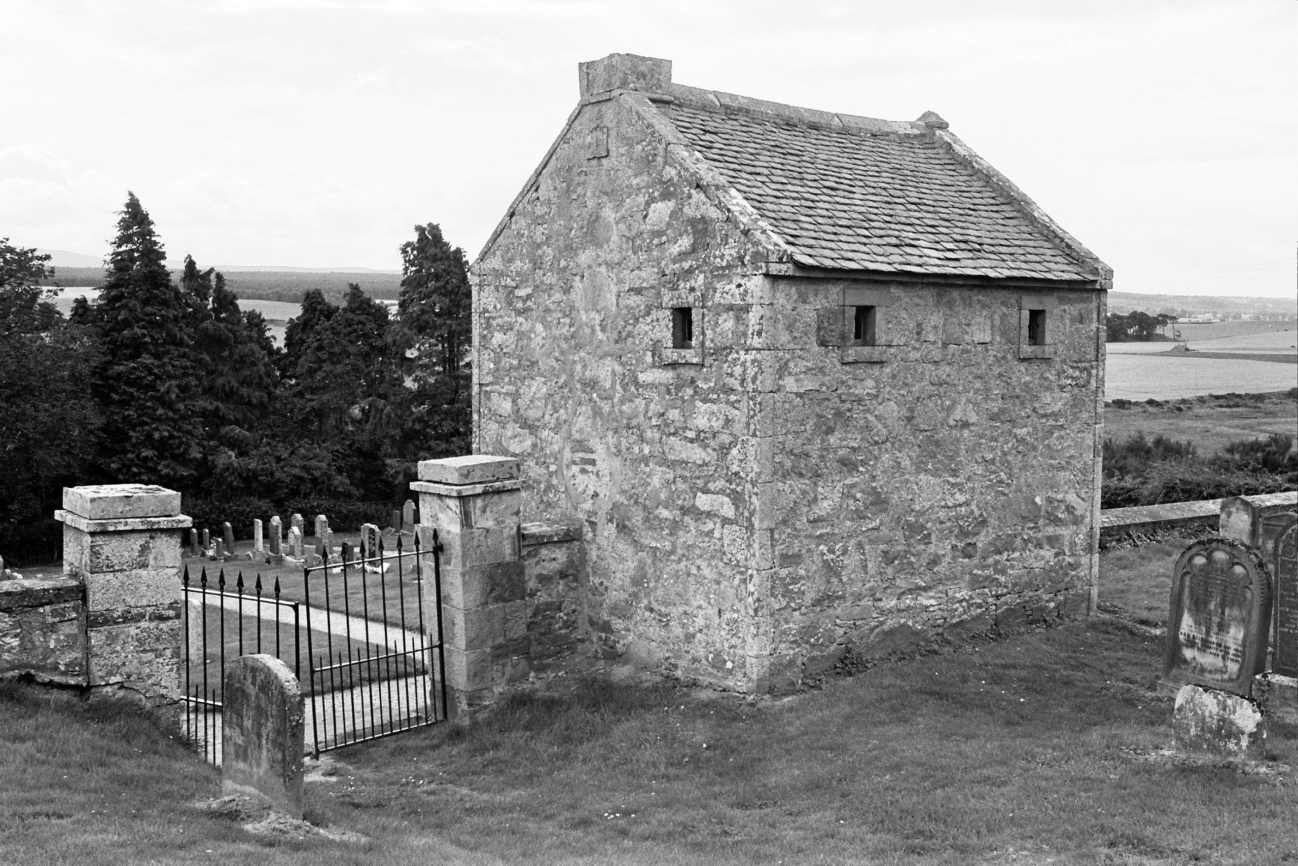 Black-and-white photograph of a small stone watch house with a pitched roof and two small square windows. The building is enclosed by a low stone wall and an iron gate, with gravestones visible in the background. Trees and open countryside stretch beyond the enclosure.