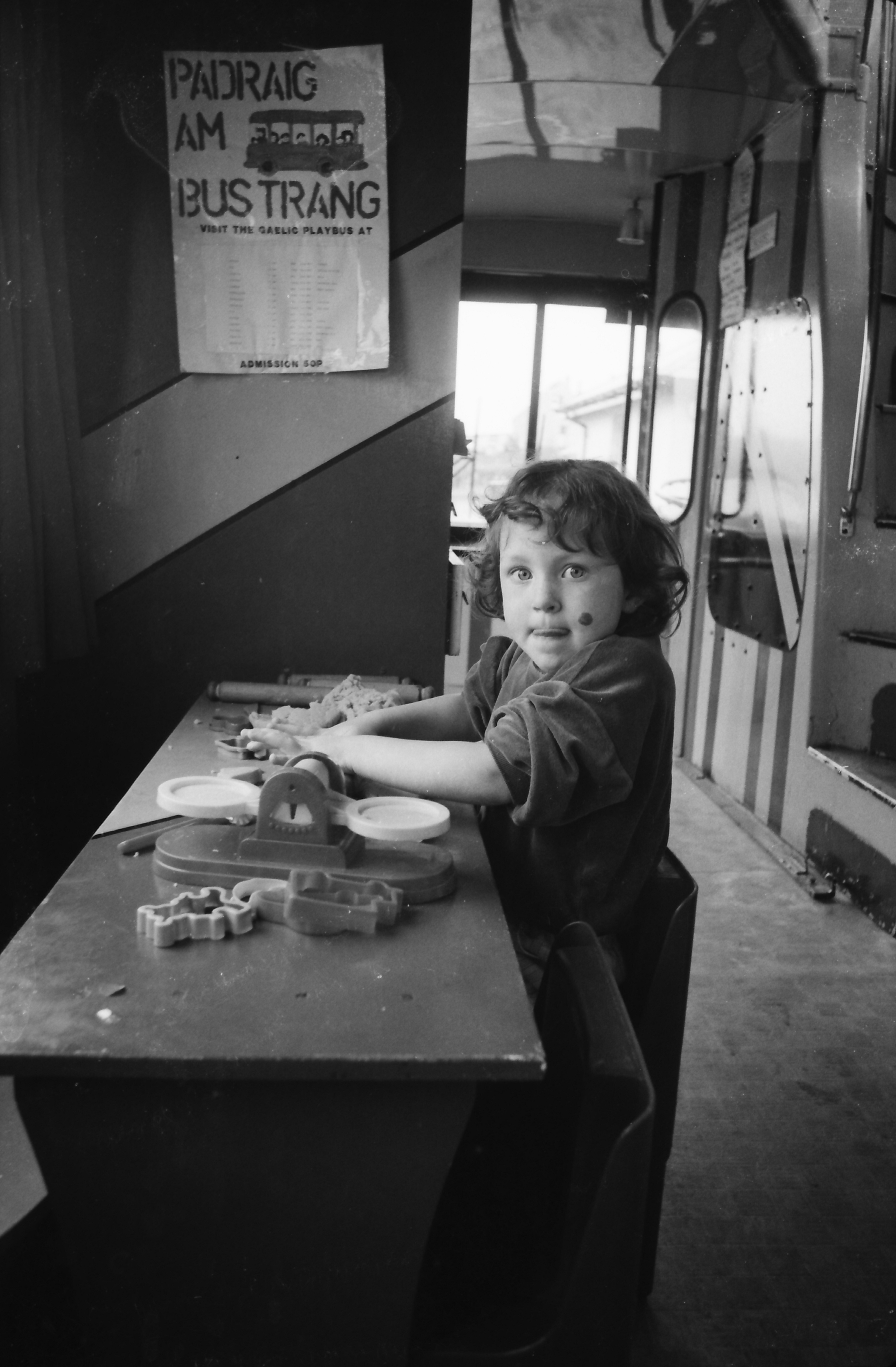 Photograph of a child taking part in activities on the Gaelic Playbus (GB3219/D263/19/27). The image shows a young child seated at a table with educational toys inside a converted bus used as a mobile Gaelic-language learning and play space. A poster reading “Pàidraig Am Bustrang” is visible on the wall. The Gaelic Playbus was developed to support early years Gaelic language learning and promote Gaelic-medium education within communities across the Highlands and Islands.