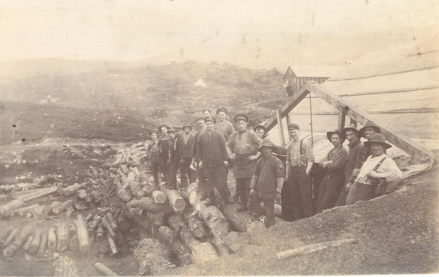 A sepia-toned historical photograph showing a group of people standing outdoors near a wooden structure with a slanted roof. The individuals are dressed in work clothes, including hats and suspenders, suggesting a labor or logging setting. In the foreground, there is a large pile of cut logs arranged along the ground. The background features a rugged, hilly landscape with sparse vegetation and a tent visible in the distance, indicating a remote or rural location. The overall scene conveys a work camp or construction site from an earlier era.